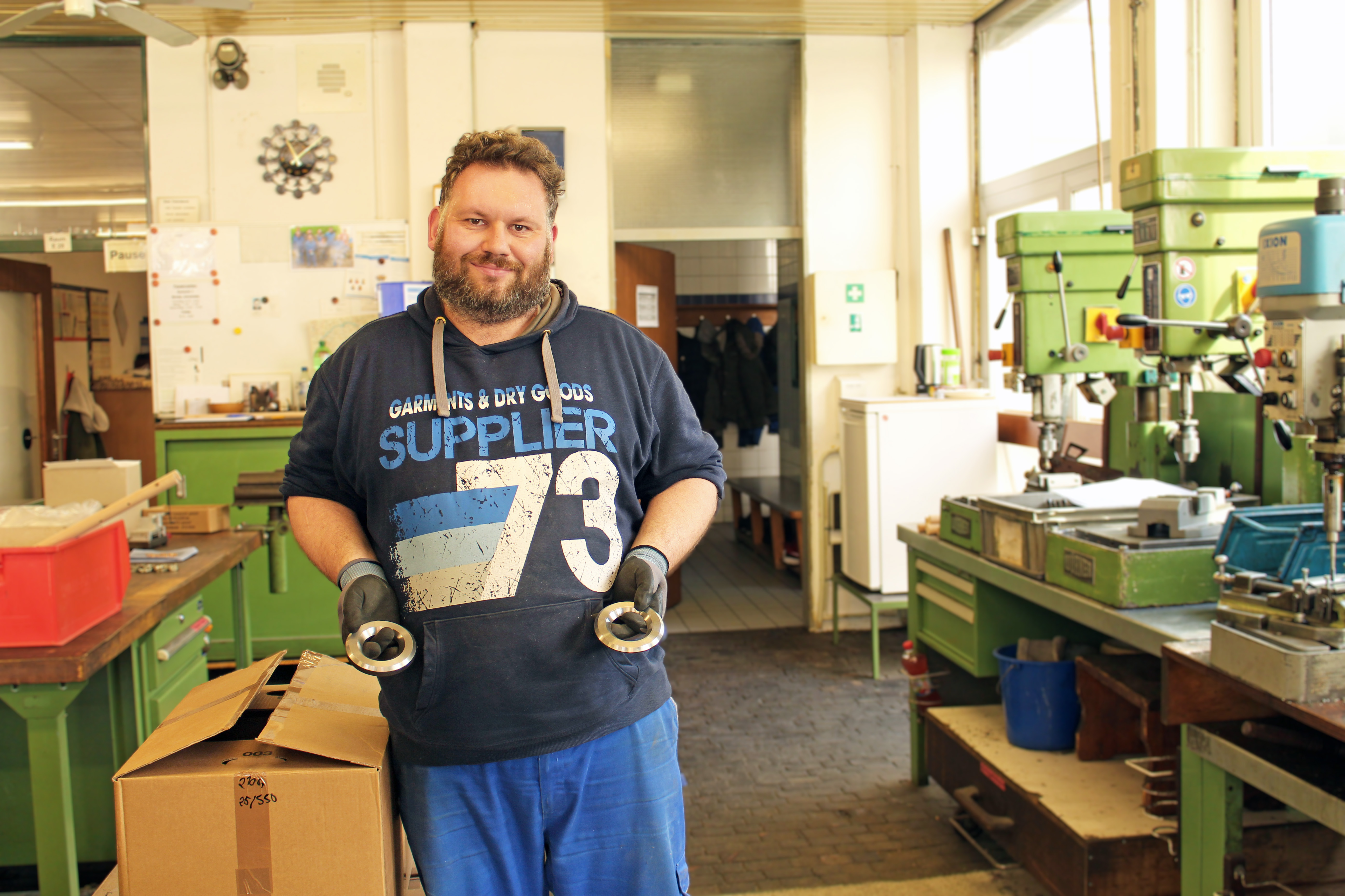 Werkstattmitarbeiter hält zwei Metallringe in den Händen, auf der rechten Seite sieht man Werkzeug und Maschinen