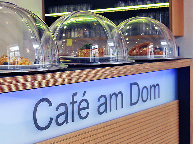 Theke im Cafe mit beleuchteter Aufschrift "Cafe am Dom". Auf der Theke stehen drei Tabletts mit Kuchen, die mit Glashauben abgedeckt sind.