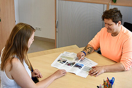 Eine Mitrabeiterin der Offenen Behindertenarbeit und einen junge Dame sitzen sich an einem Schreibtisch gegenüber. Die Mitrabeiterin deutet mit einem Stift auf eine Stelle in einem aufgeschlagenen Formular.