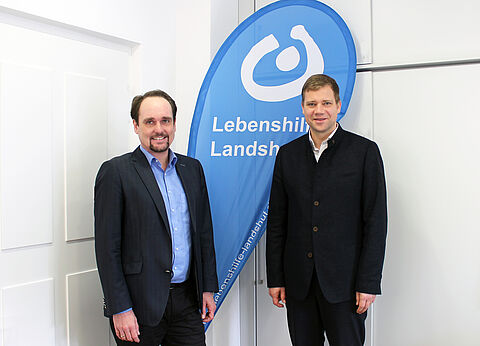 Geschaeftsfürer Johannes Fauth und Bezirktsatgsbräsident Dr. Olaf Heinrich stehen nebeneineander im Büro. Im Hintergrund ist die Fahne mit dem Logo der lebenshilfe Landshut zu sehen.