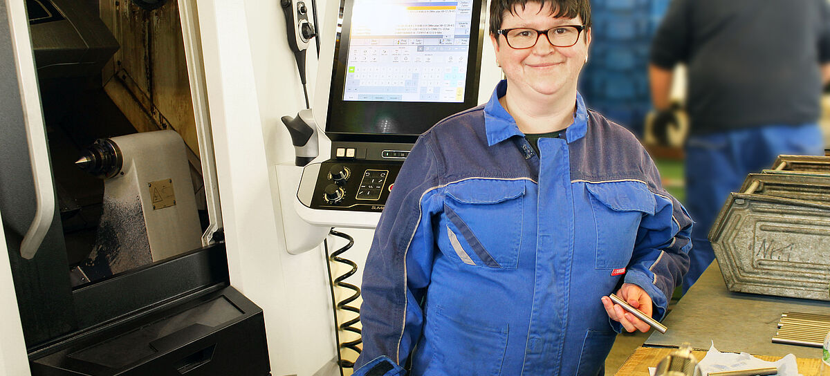 Werrkstattmitarbeiterin in Arbeitskleidung mit Werstück aus Metall in der Hand, mit großer Maschine im Hintergrund