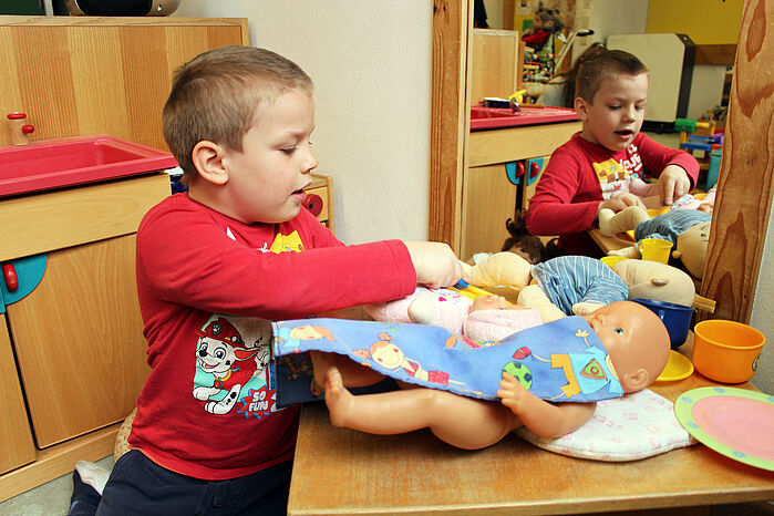 Kleiner Junge spielt Füttern mit einer Puppe, die auf einem Tisch liegt. Daneben liegt Puppengeschirr.  