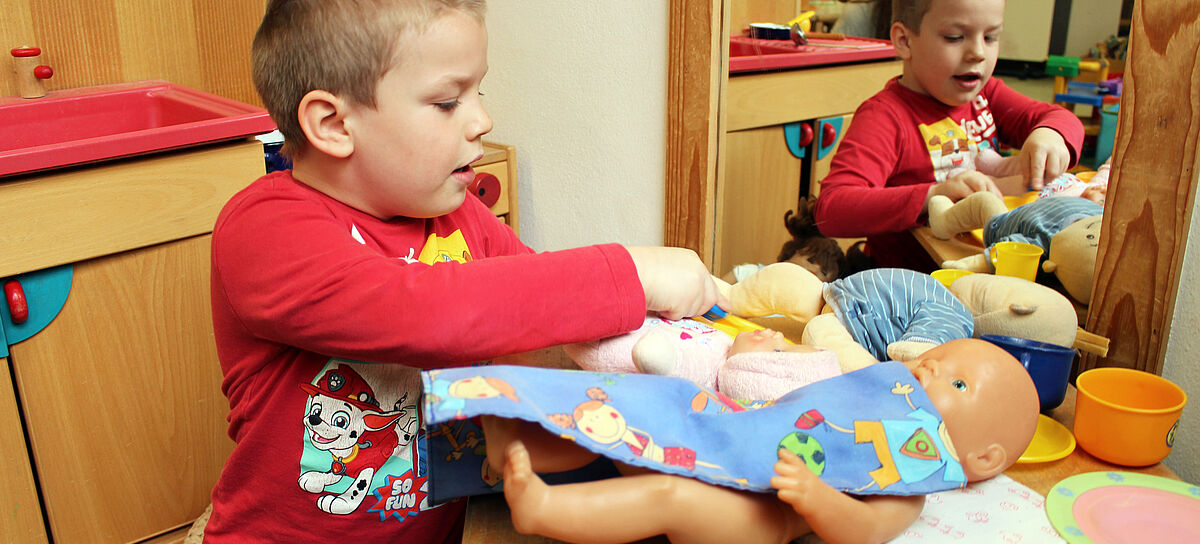 Kleiner Junge spielt Füttern mit einer Puppe, die auf einem Tisch liegt. Daneben liegt Puppengeschirr.  