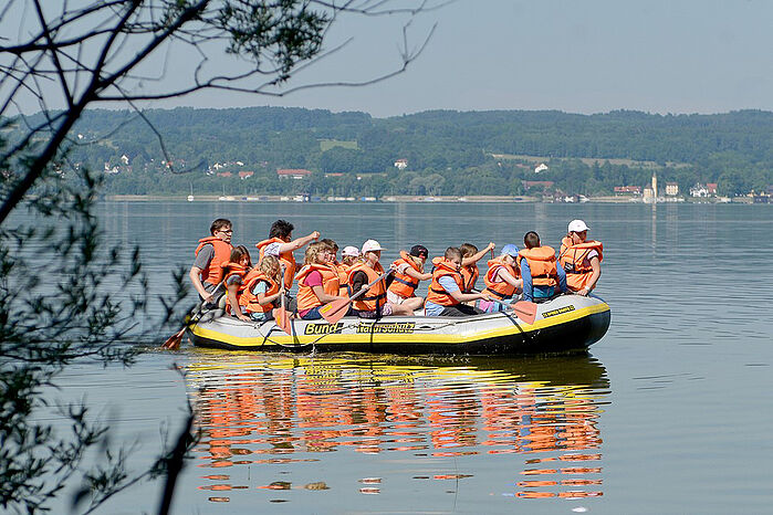 Großen Schlauchboot auf einem See mit circa 14 Personen, die Schwimmwesten tragen und Ruder halten.