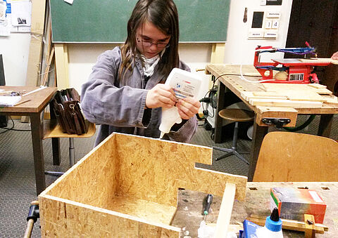 Ein Mädchen mit Arbeitskittel leimt im Werkunterricht eine Kiste zusammen