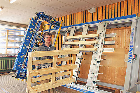 Werkstattmitarbeiter trägt eine Holzpalette, im Hintergrund ist eine Maschine zum Palettennageln zu sehen.