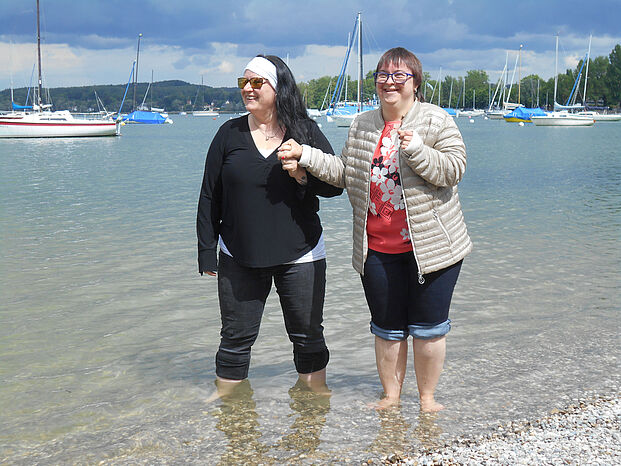 Eine Frau führt eine andere Frau an der Hand am Ufer eines Sees. Sie stenen mit den Füßen im Wasser.